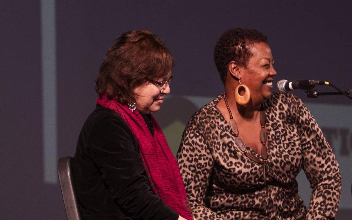 A person in brown hair, glasses, a red scarf and black cardigan sits next to a Black person wearing a leopard print shirt who is smiling in front of a microphone